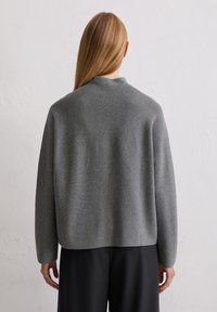 Grauer, übergroßer Strickpullover mit hohem Kragen, abgesenkten Schultern und glatter Textur, kombiniert mit schwarzen weit geschnittenen Hosen.