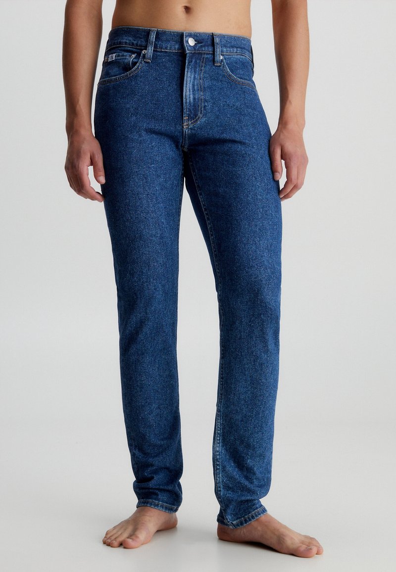 Barfota person i slimfit blå denimjeans som står mot en neutral ljus bakgrund.