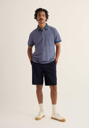 Mężczyzna stoi z rękami w kieszeniach, ubrany w niebiesko-fioletową paskowaną koszulkę polo, granatowe szorty, białe skarpetki i białe tenisówki.