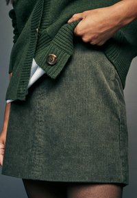 Olivgrön kordora kjol med vertikalt ribbat mönster, rak snitt och en synlig knapp på en grön stickad cardigan.