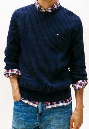 Mann trägt einen marineblauen, gerippten Pullover über einem rot-weiß-blau karierten Hemd mit hochgekrempelten Ärmeln, kombiniert mit blauen Jeans, Hände in den Taschen.