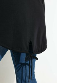 Camisa negra con dobladillo y abertura lateral, con cordón anudado sobre jeans de mezclilla azul, contra un fondo blanco.