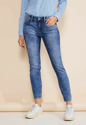 Personne portant un jean skinny bleu clair, des baskets blanches et une chemise à manches longues rayée bleue, debout avec une main dans la poche.