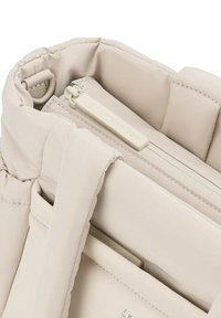 Sac à main en tissu matelassé beige avec fermeture à glissière et étiquette de logo. Présente une texture lisse et un intérieur spacieux avec des poches pour l'organisation.
