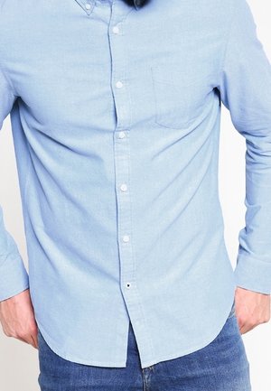 Homme portant une chemise bleu clair à boutons avec une poche poitrine et un jean bleu, debout les mains reposant près des hanches.