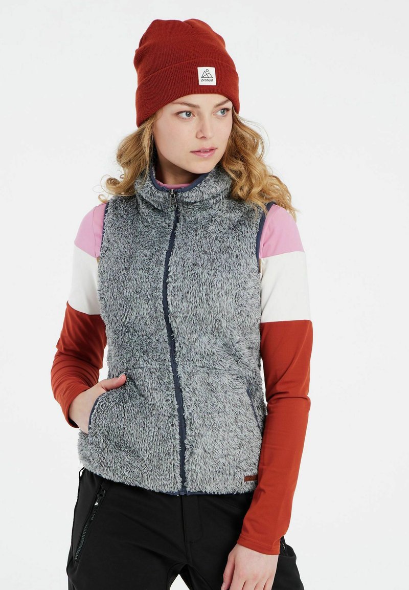 Fleece vesta v sivej farbe, s vysokým golierom a predným zipsom. Preložená s viaczlohovou košeľou s dlhým rukávom a červenou čiapkou.