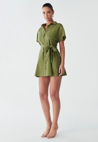 Abito camicia verde oliva con maniche corte, bottoni frontali e vita legata. Tessuto morbido con gonna a ruota e design minimalista.