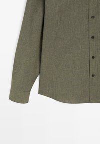 Długa koszula z długim rękawem w kolorze oliwkowej zieleni, wykonana z teksturowanego materiału. Posiada front zapinany na czarne guziki oraz zaokrąglony dół.