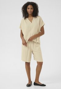 Haut beige avec un col en V et des manches courtes associé à un short beige de longueur genou. Modèle portant des ballerines noires, se tenant contre un fond clair.