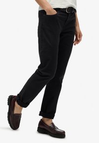 Pantalones de algodón negros con corte recto, puños enrollados y bolsillos delanteros, combinados con mocasines burdeos oscuros con detalles texturizados.