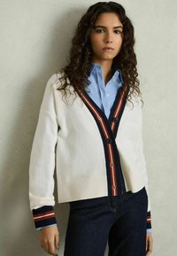 Vitt cardigan med marinblå och röda ränder, med två knappar och ribbade muddar. Bärs över en ljusblå skjorta med knappar. Jeansen är av denim.