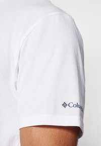 Valkoinen puuvillainen lyhythihainen paita, jossa on suora helma. Vasemmassa hihassa on tummansinisellä värillä "Columbia"-logo. Pehmeä pinta, yksinkertainen muotoilu.