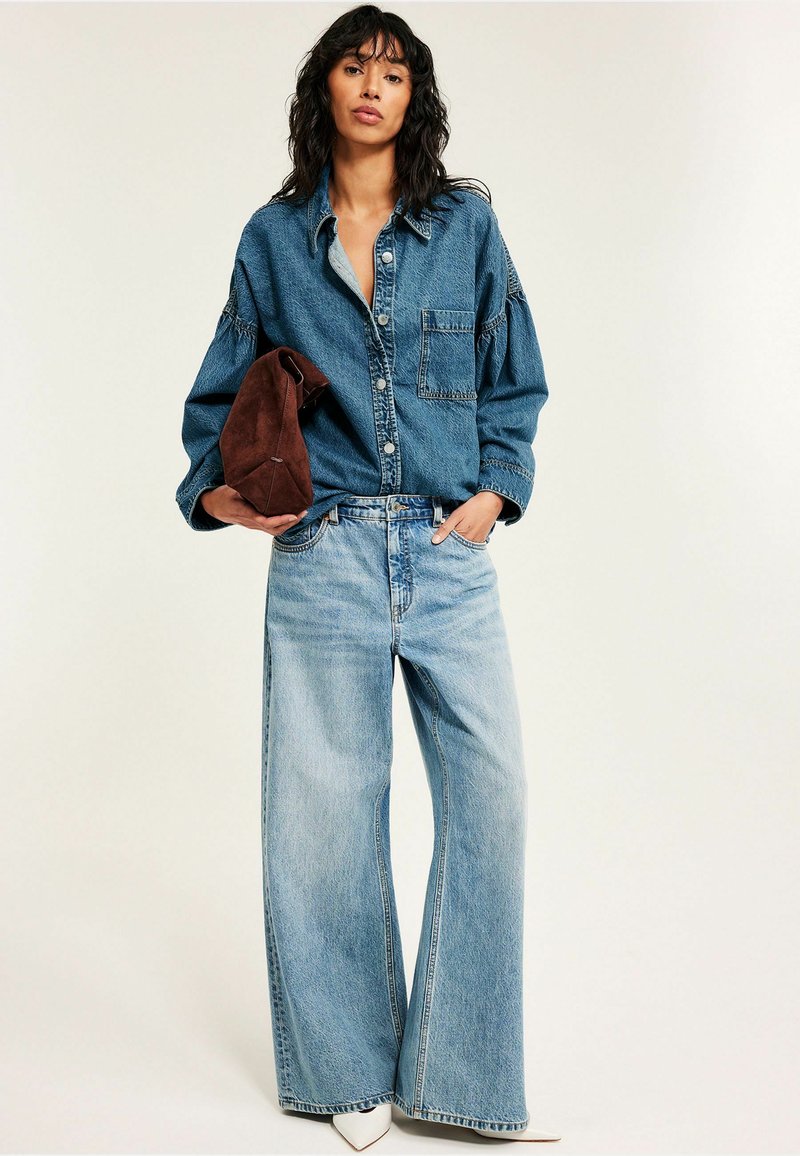 Woman wearing oversized denim shirt and wide-leg jeans, holding a brown suede bag, standing against a plain light background.