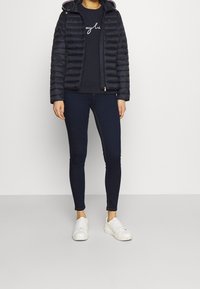 Chaqueta acolchada azul oscura con capucha, combinada con un sudadera navy y jeans oscuros. Zapatillas blancas completan el look. Texturas suaves en todo.
