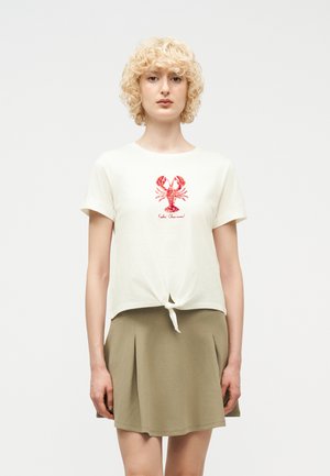 Blondynka o kręconych włosach nosi białą koszulkę z czerwonym nadrukiem homara i zawiązanym supełkiem, w połączeniu z beżową plisowaną spódnicą.