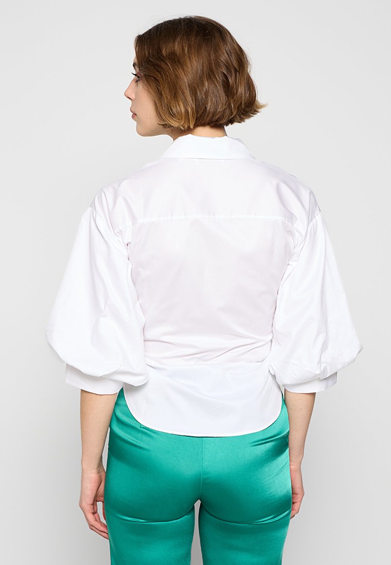 Weißes Oberteil mit Puffärmeln, glatter Textur und taillierter Passform, kombiniert mit grünen Satin-Hosen mit glänzendem Finish.