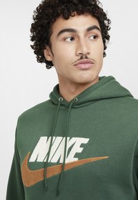 Felpa verde con tasca a marsupio frontale, caratterizzata da un logo Nike bianco e arancione. Materiale in cotone con una texture morbida e cappuccio con cordino.