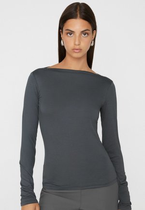 Frau mit langen dunklen Haaren, die silberne Creolen trägt und ein eng anliegendes dunkelgraues Langarmshirt vor einem einfarbigen hellen Hintergrund.