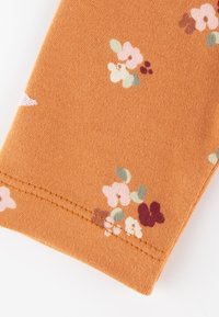 Orangener Stoff mit glatter Textur, der kleine pastellfarbene Blumenmuster in Rosa, Grün und Kastanienbraun zeigt, mit sichtbaren Nähten am Rand.