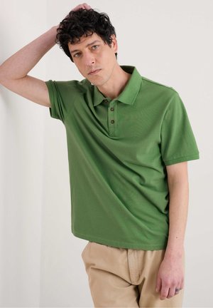 Camisa polo verde de manga corta, con un cuello clásico y tres botones marrones. Combinada con pantalones beiges para un contraste neutro.