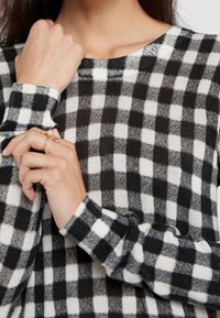 Schwarz-weißer karierten Pullover mit langen Ärmeln, rundem Ausschnitt und kontrastierenden Bündchen. Das Material scheint weich mit einer strukturierten Oberfläche zu sein.
