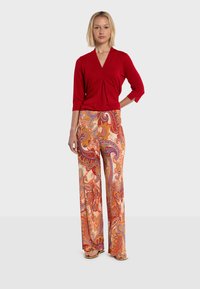 Blouse rouge à manches longues avec un devant plissé, associée à un pantalon beige arborant un motif paisley coloré, une coupe large et une texture douce.