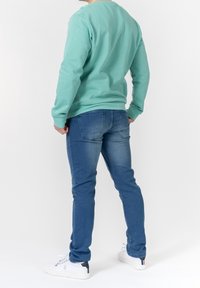Man wearing a mint green sweatshirt, blue jeans, and white sneakers, standing facing away with hands in pockets against white background.