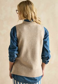 Beige gestrickte Weste mit einem gerippten Saum, über einem blauen Jeanshemd getragen. Merkmale einen Rundhalsausschnitt und eine weiche Textur, die einen schichtartigen Look kreiert.