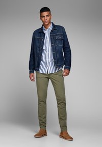 Jeansjakke, stribet skjorte med knapper, olivengrønne bukser og brune ankelstøvler. Funktioner inkluderer kontrastsyninger og en skræddersyet pasform.
