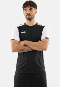 Zwart sportshirt met korte mouwen, witte mouwen en grijze accenten. Heeft het "JAKO" logo op de bovenkant van de linkerkant van de borst. Lichtgewicht stof.