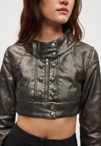 Μεταλλικό κοντό μπουφάν με ψηλό γιακά, ασημένιο φερμουάρ και διακοσμητικά κουμπιά· υφή με αμυδρή γυαλάδα και εφαρμοστή γραμμή.