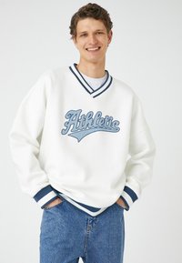 Sudadera blanca con rayas marinas en el cuello y los puños; presenta texto bordado en azul "Athletic" en el pecho; corte relajado.