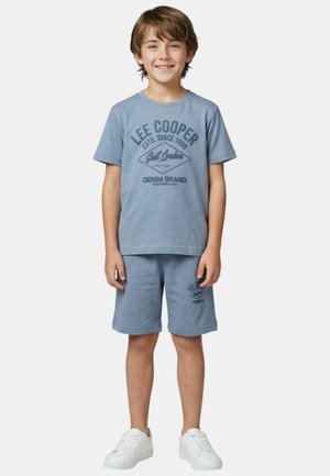 Garçon debout souriant, portant un t-shirt bleu de la marque Lee Cooper et un short, avec des baskets blanches, sur un fond uni.