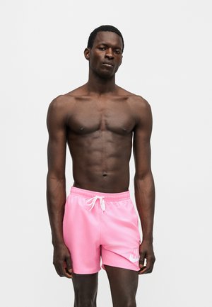 Homme musclé torse nu à la peau foncée portant un short de sport rose vif avec un cordon blanc, debout devant un fond gris clair uni.