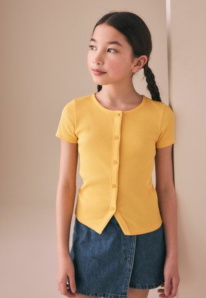Mädchen mit geflochtenen Haaren, das ein kurzärmeliges gelbes Hemd und einen blauen Jeansrock trägt, schaut seitlich vor neutralem Hintergrund.
