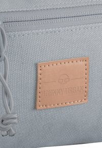 Lichtblauwe stofzak met een gestructureerd oppervlak, voorzien van een gestikt bruin leren label met reliëftekst en een ritssluiting.