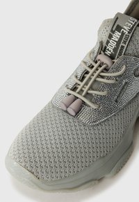 Grå sportsko med mesh-overdel, struktureret detaljer, elastiske snørebånd og en markant blomst med refleksaccenter på tungen.
