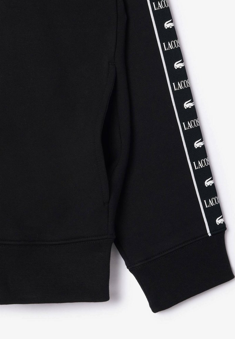 Schwarzer Pullover mit umgenähtem Saum und einer Seitentasche, versehen mit einem vertikalen weißen Streifen, der mit wiederholten Lacoste-Logos geschmückt ist.