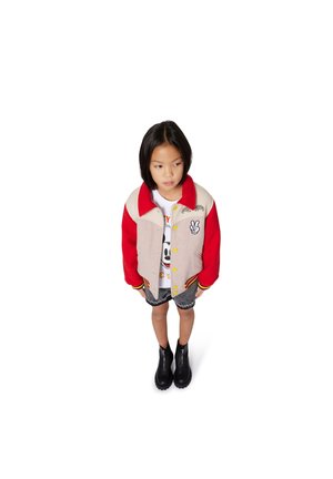 Giacca varsity rossa e beige con bottoni gialli, t-shirt grafica, pantaloni corti in denim e stivaletti neri alla caviglia. Il bambino si trova in piedi con le mani sui fianchi.