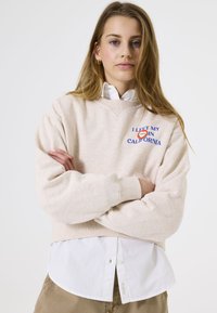 Beige sweatshirt met een ronde hals, geborduurde tekst "IK HEB MIJN IN CALIFORNIA GELATEN," gelaagd over een witte overhemdd, casual pasvorm.