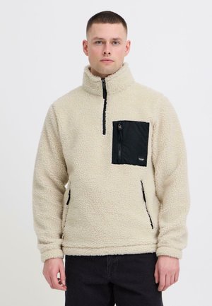 Homme portant un pull en polaire crème avec une poche poitrine zippée noire et des poches latérales zippées noires, debout devant un fond uni.