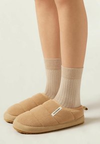 Beige Hausschuhe mit gerippter Textur, runder Zehenpartie und einem Etikett mit der Aufschrift "CALZEDONIA". Getragen mit hellen, gerippten, metallischen Socken.