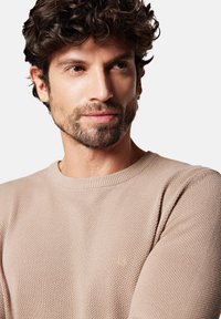 Beżowy sweter dziany z okrągłym dekoltem i teksturowanym materiałem. Posiada subtelną markę na piersi, dopasowane rękawy i gładkie wykończenie.