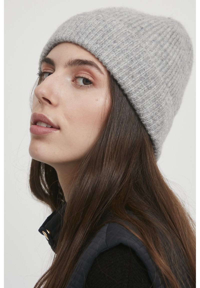 Beanie grigio a coste, texture morbida, forma aderente, indossato leggermente di lato. Gilet nero con hardware dorato visibile al colletto.