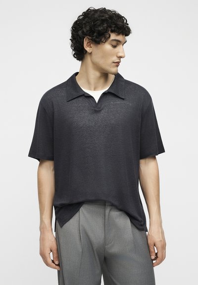 Νέος άνδρας με κοντά σγουρά μαλλιά, ντυμένος με σκούρο χαλαρό πόλο με κοντά μανίκια και ανοιχτό γκρι παντελόνια, κοιτάζοντας στο πλάι.