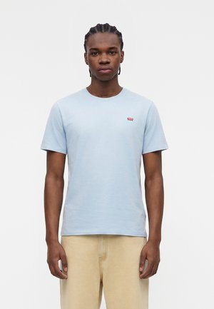 Νέος άνδρας με πλεξούδες, φορώντας ανοιχτό μπλε μπλουζάκι με κοντά μανίκια και μπεζ παντελόνι, στέκεται με χαλαρά χέρια μπροστά σε λευκό φόντο.