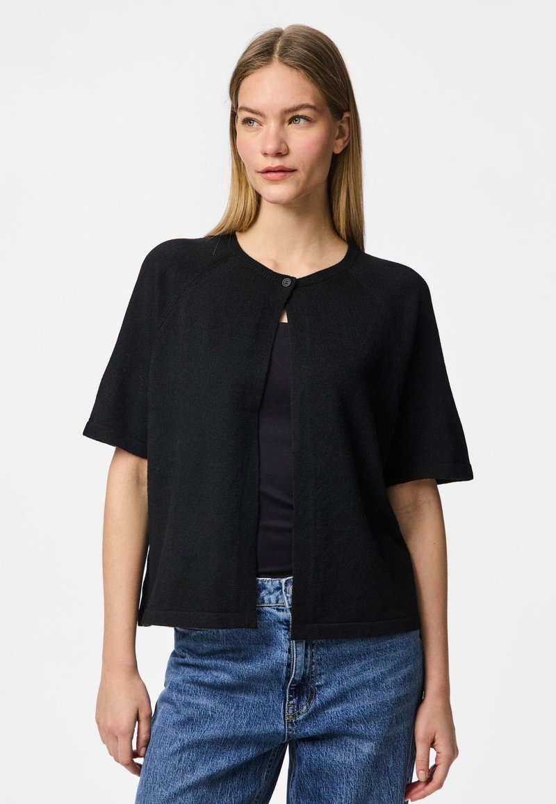 Cardigan preto de mangas curtas com gola redonda e fecho de botão único. Tecido leve e texturizado, combinado com uma blusa preta e jeans azuis.