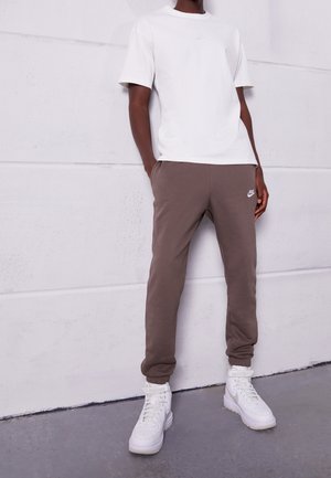 Person trägt weißes Nike-T-Shirt, braune Nike-Jogginghose und weiße High-Top-Sneaker, steht mit den Händen in den Taschen vor einer weißen Wand.