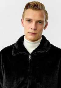 Zwarte nepbont jas met een hoge kraag en ritssluiting, gedragen over een witte coltrui. Dankzij de soepele textuur heeft het een luxueuze uitstraling.