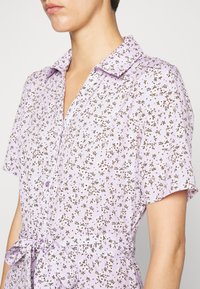 Vestido camiseiro lilás com mangas curtas, frente com botões e padrão floral em castanho escuro e branco. Inclui detalhe na cintura amarrada.